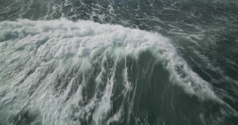 A dramatic slow-motion aerial follows a powerful ocean wave as it forms, crests, and crashes over a rocky shoreline