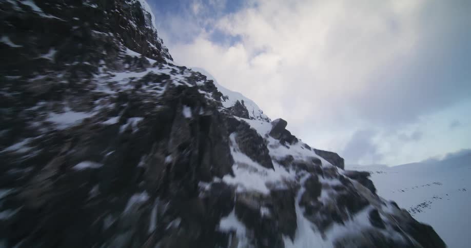 A dramatic aerial view ascending a rugged, snow-covered mountain face with frozen waterfalls under a cloudy sky