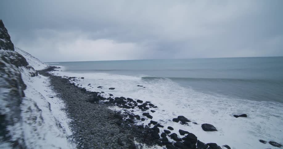 A dramatic aerial shot flies along a snow-covered cliff, revealing a rocky coastline with powerful waves crashing ashore on a cold, overcast day