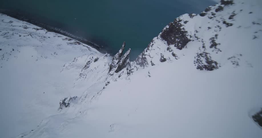 An FPV drone soars down the side of a massive snow-covered mountain ridge overlooking the icy sea below