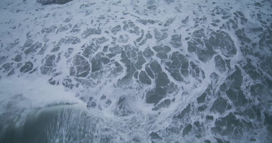 An overhead slow-motion shot follows a powerful ocean wave as it rolls and breaks into churning white seafoam