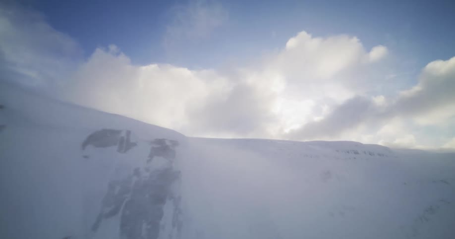 An aerial shot flies backwards over a majestic snow-covered mountain as strong winds blow snow across the desolate landscape