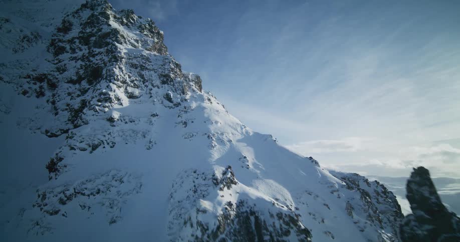 An aerial view flies past a rugged, snow-covered mountain peak against a vast blue sky