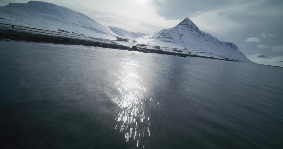 A dramatic FPV drone flight speeds over the cold ocean towards the snow-covered base of Iceland's Kirkjufell mountain as the sun glints off the water