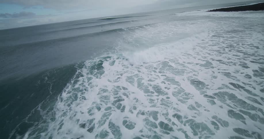 A dramatic slow-motion aerial follows a powerful ocean wave breaking as a seagull flies over surfers in the cold water near a snowy shore