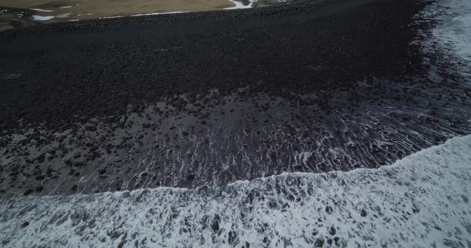 A dramatic aerial shot follows white sea foam as it recedes from a black volcanic pebble beach