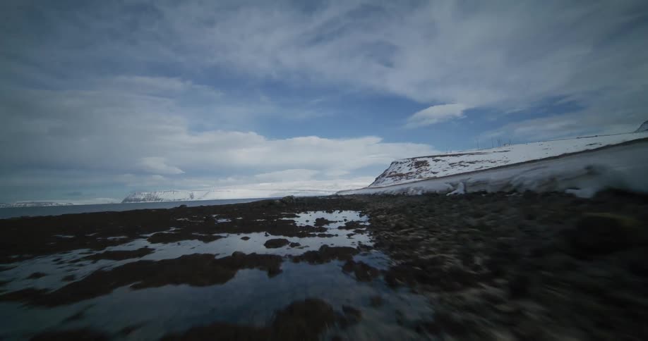 Dynamic FPV drone footage soars low and fast along a snowy bank and a rocky shoreline in a vast Nordic coastal landscape
