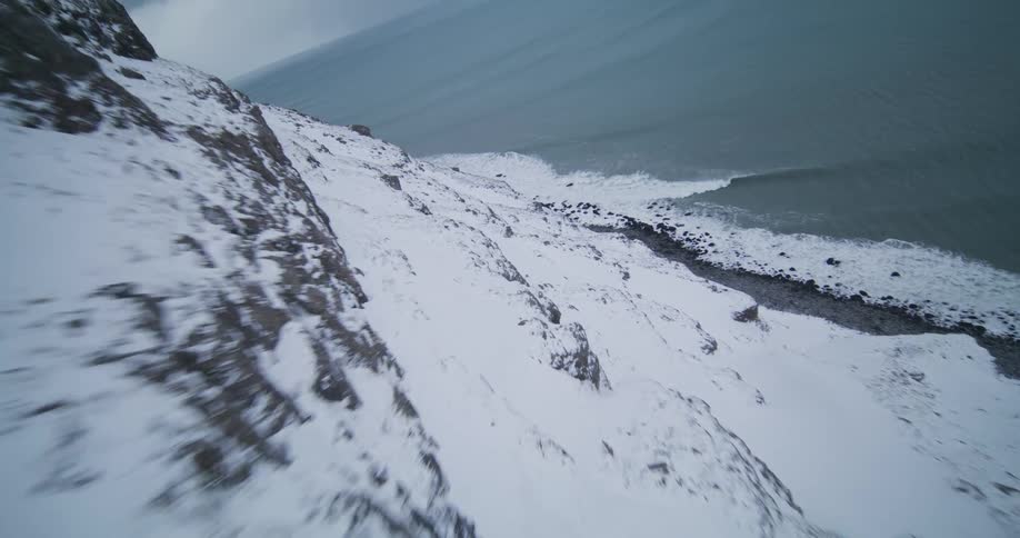 A thrilling FPV drone flight races along a steep, snow-covered cliffside overlooking a cold, choppy ocean
