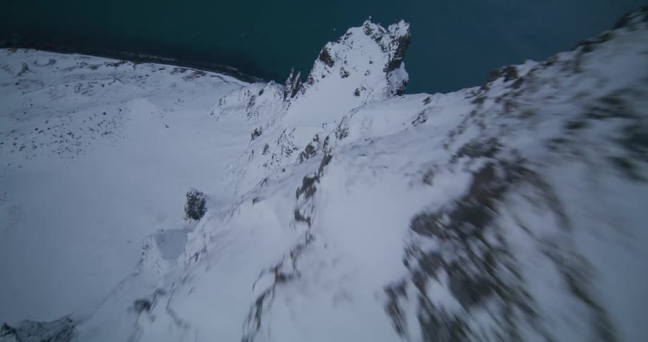 An exhilarating FPV drone flight soars over and dives down a rugged, snow-covered mountain ridge along a dramatic coastal landscape