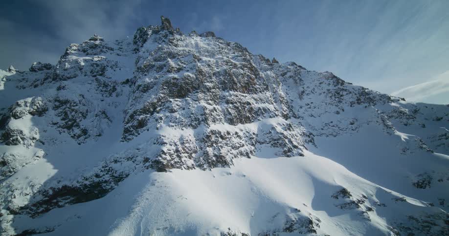 An epic aerial ascent reveals the immense scale of a rugged, snow-covered mountain peak against a blue sky
