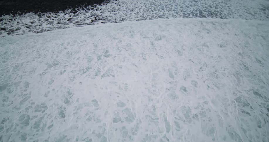 A powerful ocean wave crashes in slow motion, its white foam spreading across a black stone beach in a dramatic aerial view