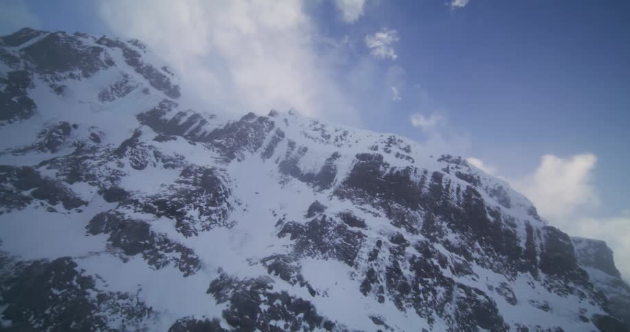 An aerial shot slowly ascends a snow-covered, rocky mountain face during a heavy blizzard