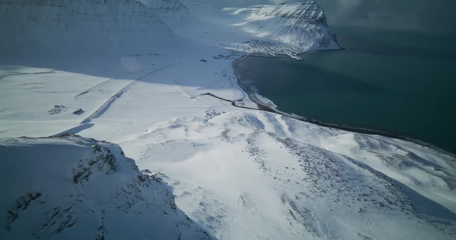 An epic aerial drone shot soars over a vast, snow-covered mountain range to reveal a remote coastal town by a fjord