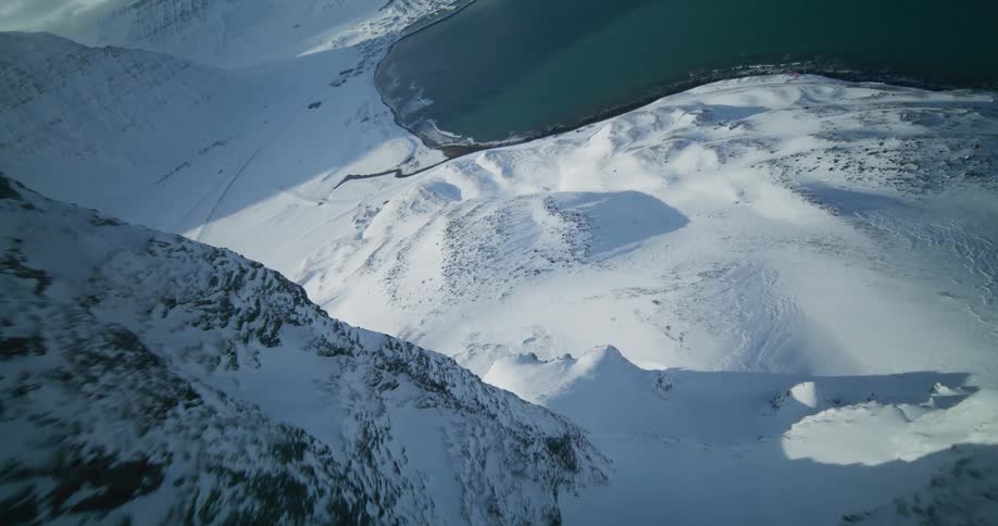 A breathtaking aerial shot flies alongside a sunlit, snow-covered mountain peak, revealing a vast frozen coastal landscape below