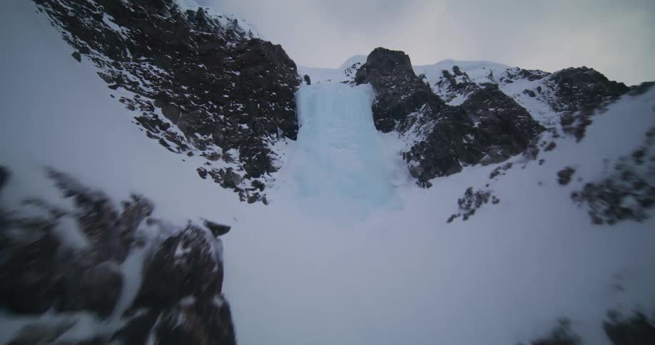 A stunning low-angle aerial shot ascends a rugged, snow-covered mountain, flying through a ravine, past a frozen waterfall, and up to the cloudy peak