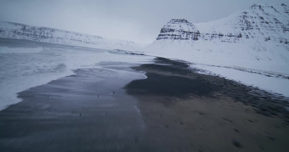 A dynamic low-flying aerial shot glides along a desolate, snow-covered shoreline as icy waves meet the black pebble beach