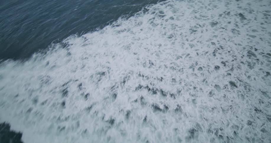 A flock of birds flies low over the white-capped ocean waves as they roll towards a dark, rocky shore in this dramatic aerial shot