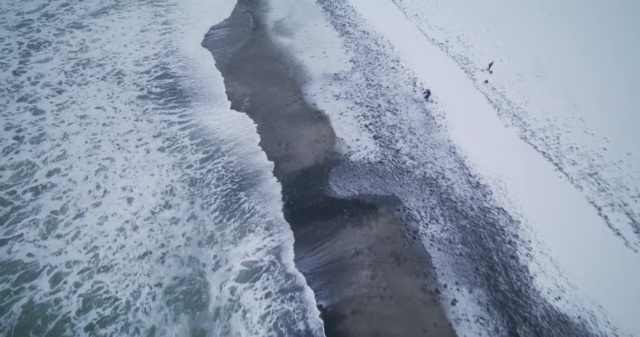 A dramatic aerial shot captures the stark beauty of a snow-covered beach as powerful ocean waves crash ashore