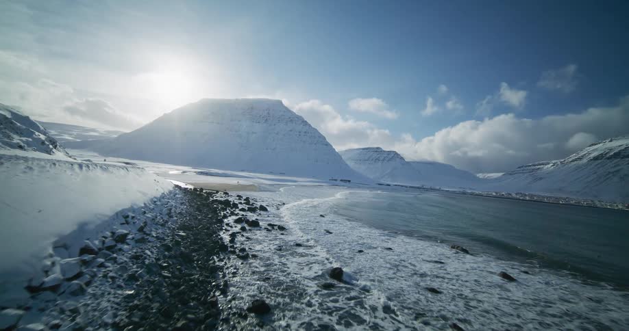 A stunning aerial view of a sunlit, snow-covered mountain landscape meeting the icy ocean waves along a rugged coastline