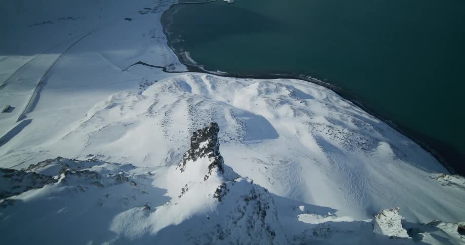 A breathtaking aerial view soars over majestic, snow-covered mountain peaks towards a remote coastal town nestled by a tranquil bay