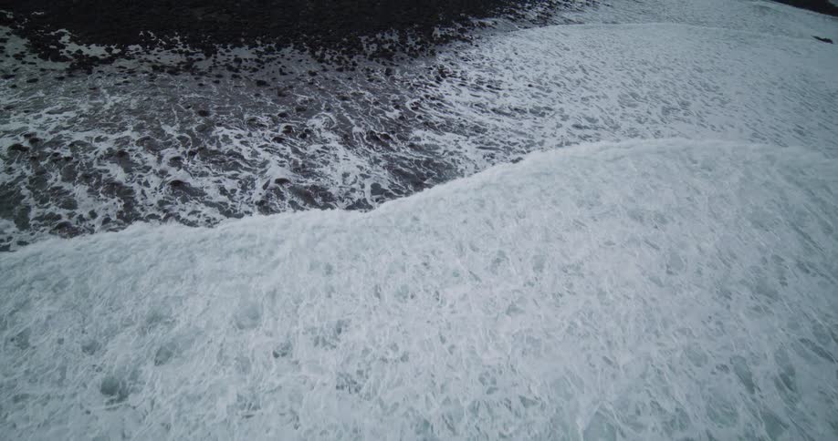A slow aerial view captures the contrast of white ocean foam against a dark, rocky coastline