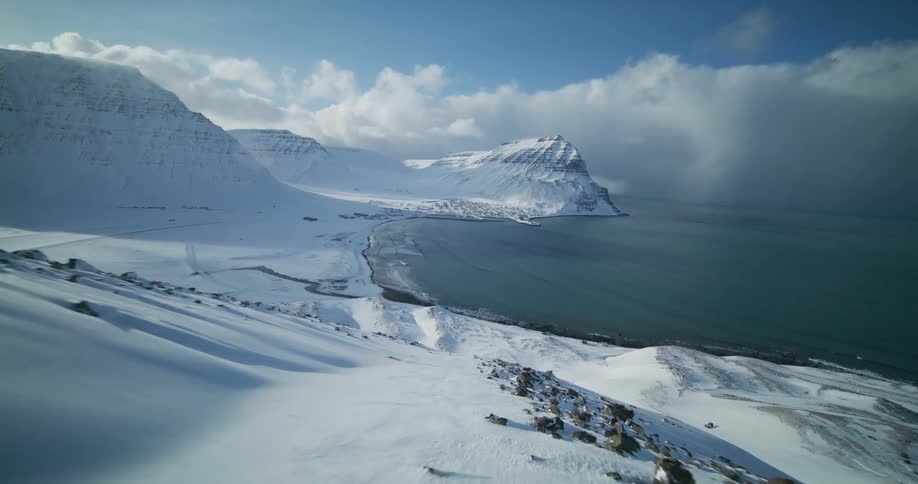 A stunning aerial view flies backward over a vast, snow-covered landscape revealing a coastal town nestled between mountains and the sea