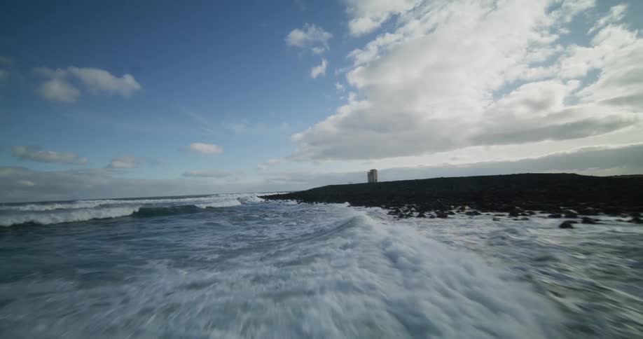 A low aerial flight skims over the ocean waves as they crash onto a dramatic, rocky shoreline towards a solitary tower
