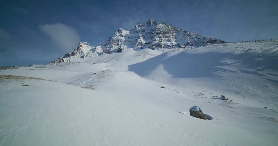 An aerial view glides over a vast, snow-covered mountain landscape toward a rugged peak on a clear winter day