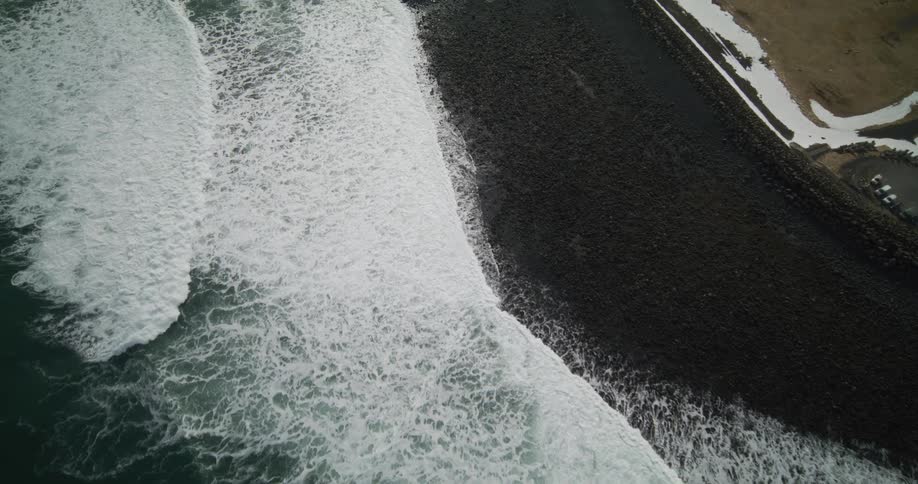A dramatic top-down aerial view captures powerful white ocean waves slowly rolling onto a black sand beach