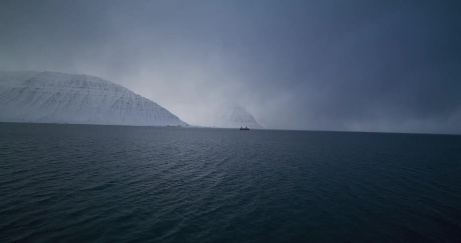 A dramatic aerial shot flies from a snowy shoreline with small houses out over a vast, dark fjord surrounded by snow-covered mountains