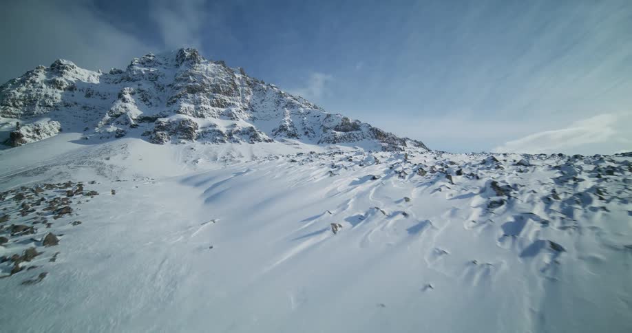 A majestic aerial flight over a vast, rocky, snow-covered landscape towards a towering mountain peak on a sunny winter day