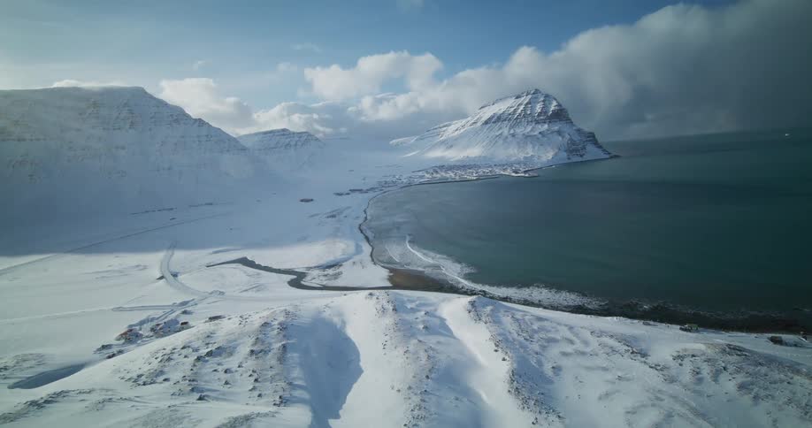 An epic aerial flight over a vast, snow-covered landscape reveals a remote coastal village nestled between majestic mountains and the ocean