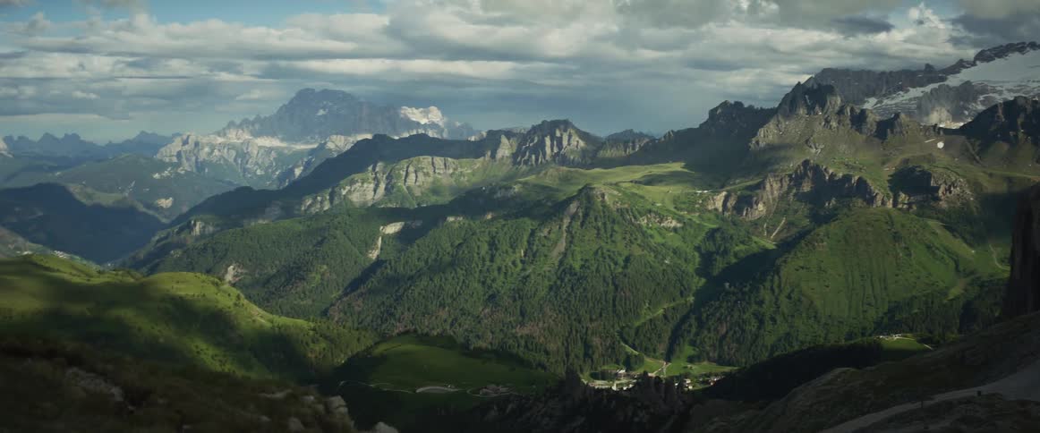 A majestic panoramic view of a sprawling green mountain valley with sunlit peaks under a dramatic cloudy sky