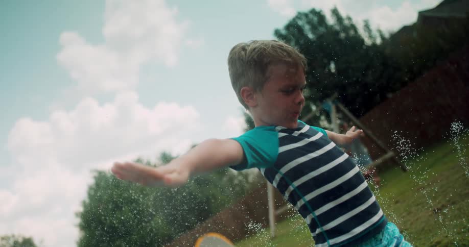 A boy is spinning in the yard over the sprinklers
