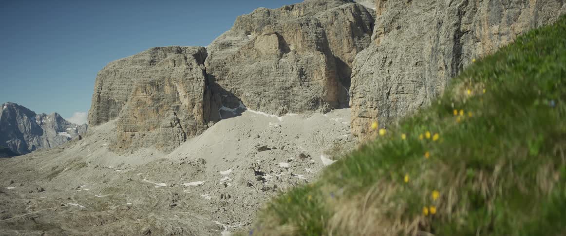 The stark beauty of a rugged, rocky mountain is softened by a grassy, wildflower-covered hillside in the foreground on a clear day