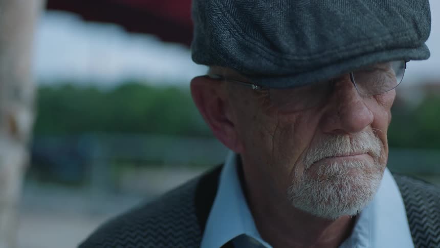 An elderly caucasian men wearing reading glasses and a hat and a frown on its face