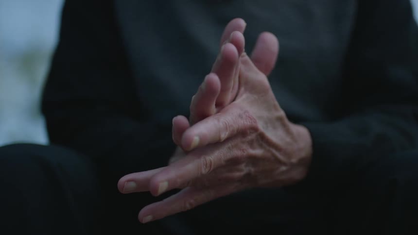 An elderly person wearing all black rubbing their hands together while sitting