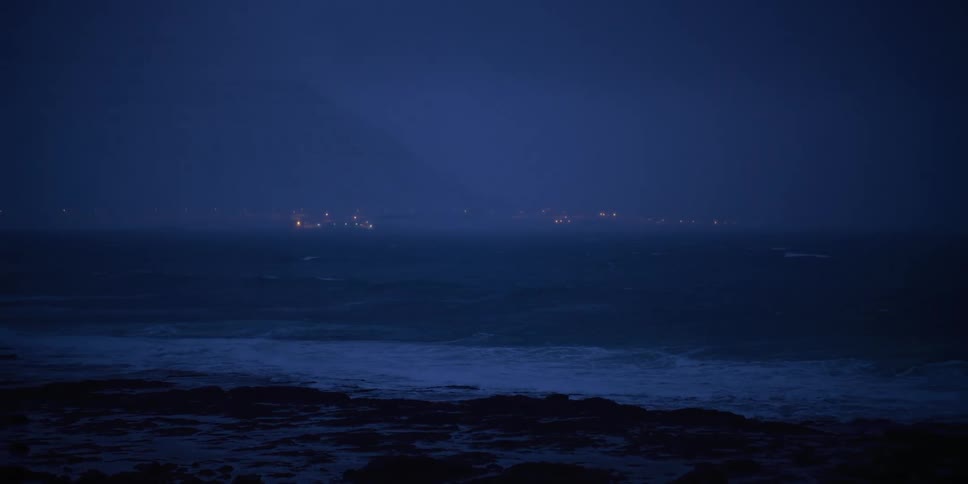 Dark, moody waves crash onto a rocky shore at night with the lights of a distant coastal town visible on the horizon