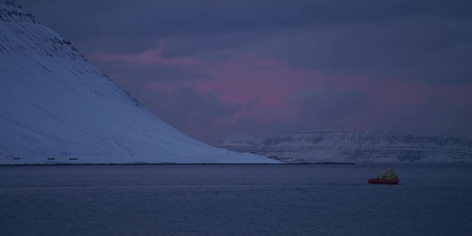 A small red boat floats on the calm waters of a fjord surrounded by snow-covered mountains at dusk