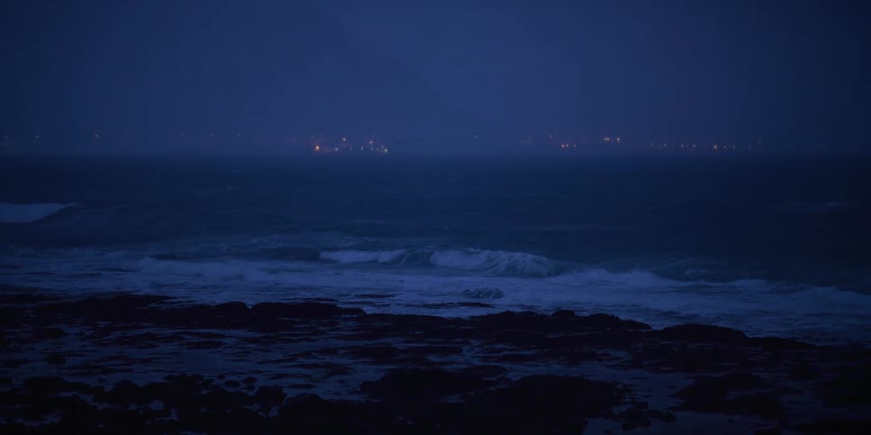 Stormy ocean waves crash against a rocky shore at night with the lights of a distant city glowing on the horizon