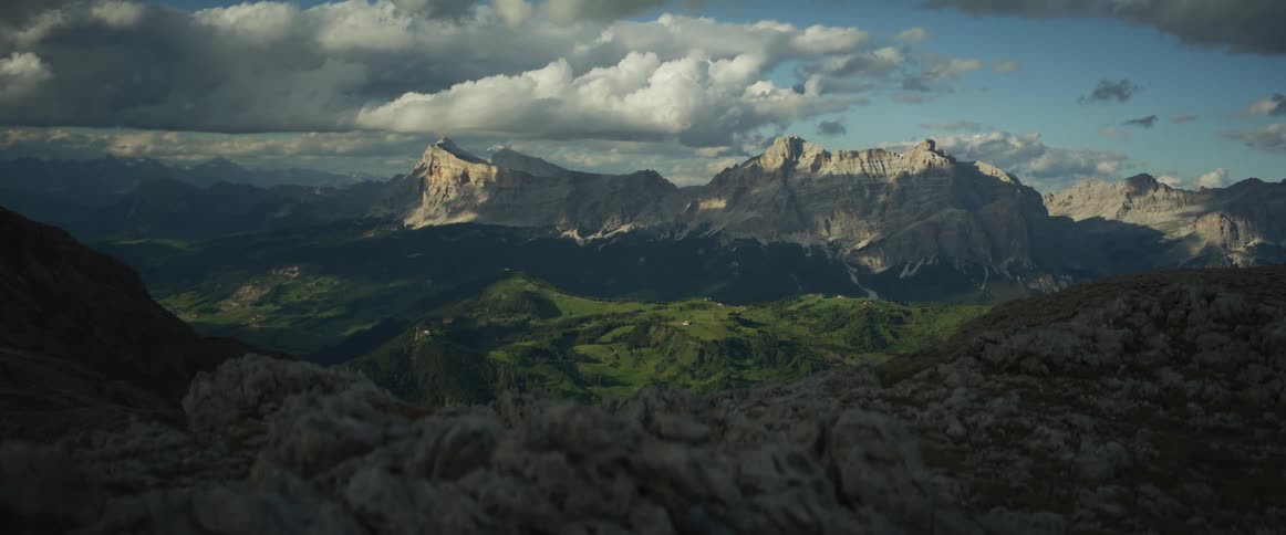 A majestic mountain range towers over a lush green valley as dramatic clouds cast moving shadows in the late afternoon sun