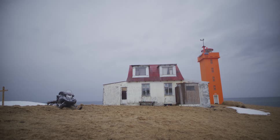 An abandoned house and a bright orange lighthouse sit on a desolate, grassy hill by the sea with a snowmobile parked nearby