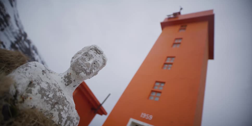 A weathered white abstract statue sits in the foreground with a bold orange lighthouse tower rising into the overcast sky behind it