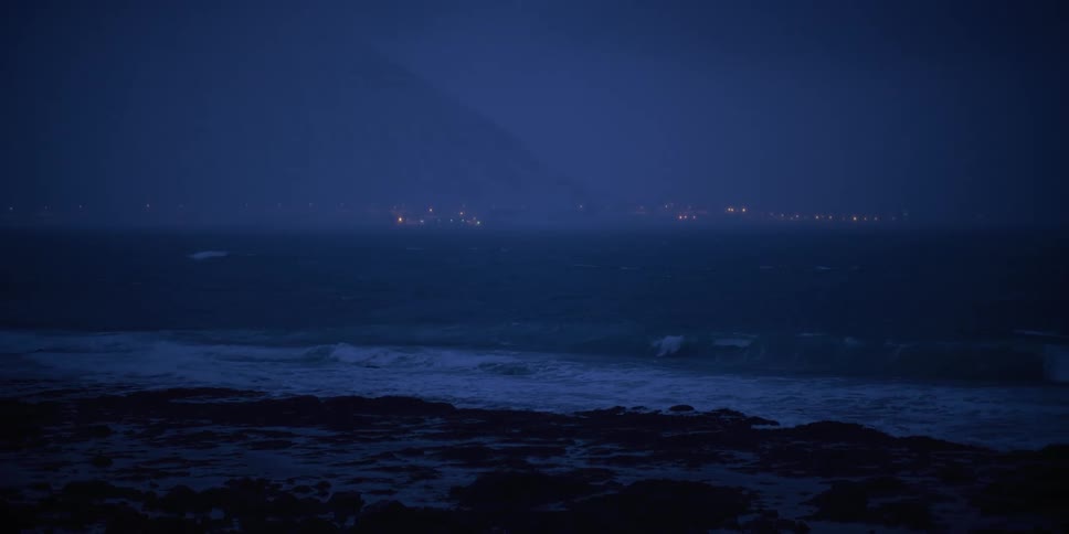 Slow-motion view of ocean waves crashing on a rocky shore during a dark, moody night with distant city lights on the horizon