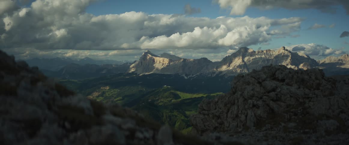 A stunning view of a vast mountain range with sunlit peaks and green valleys under a dramatic, cloudy sky