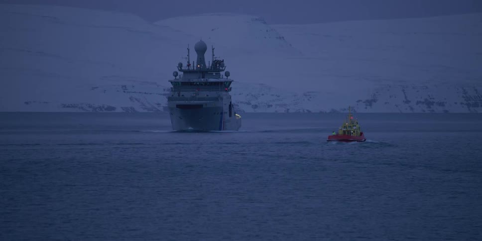 A large naval patrol ship is escorted by a small tugboat through icy waters at dusk against a backdrop of snow-covered mountains
