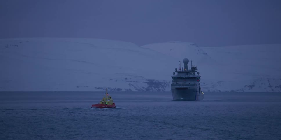 A tugboat guides a large vessel through a tranquil arctic fjord at twilight against a backdrop of snowy mountains