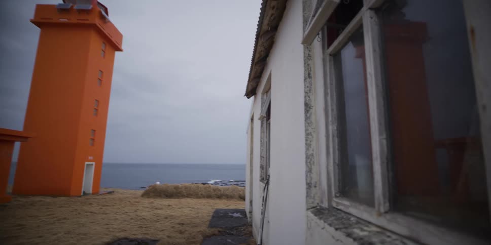 A slow reveal shot of a bright orange lighthouse standing on a desolate coastline next to a weathered and abandoned building