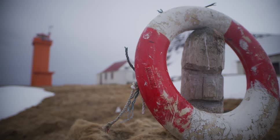 A weathered red and white lifebuoy rests on a wooden post in a snowy coastal landscape with an orange lighthouse in the background
