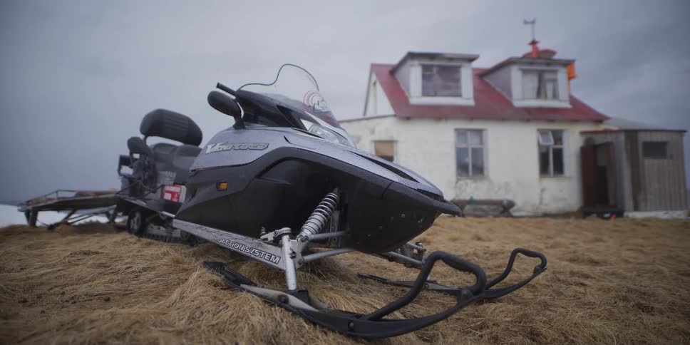 A snowmobile rests on a patch of dry grass in front of a rustic farmhouse under a grey, overcast sky